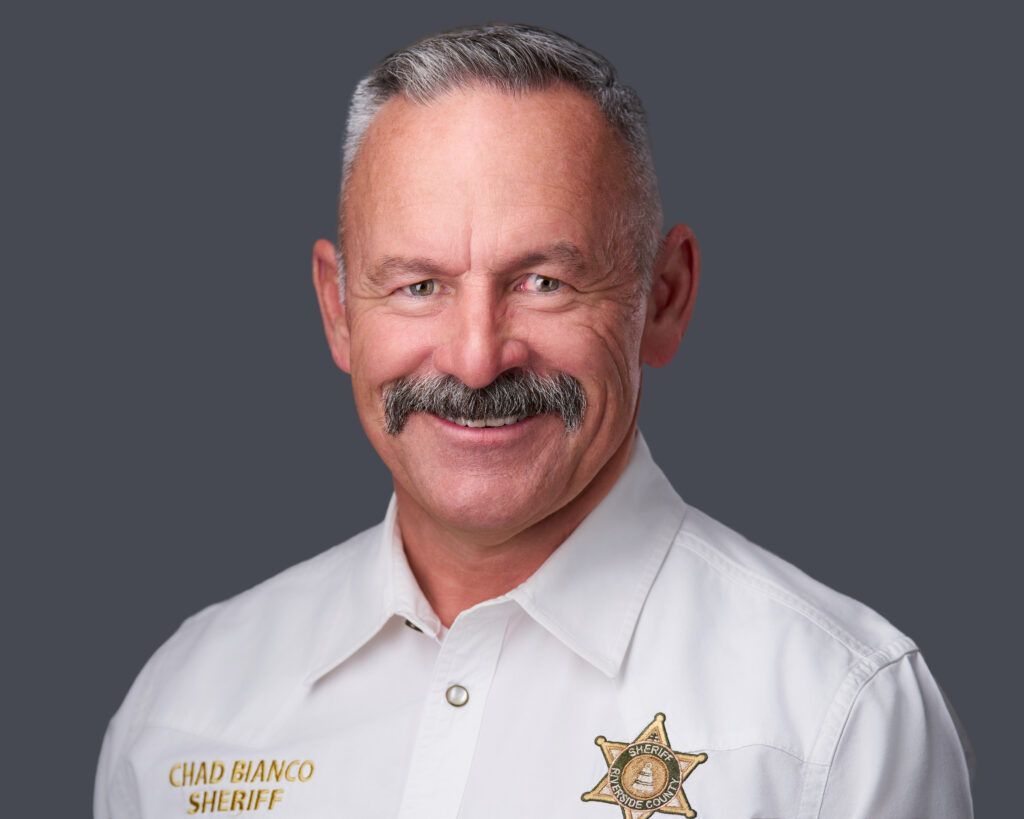 Sheriff Chad Bianco, wearing a white uniform shirt with a badge, smiling, against a gray background.
