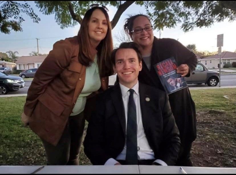 A man in a suit sits outdoors with two women. They smile, posing together. The woman on the right holds a book.