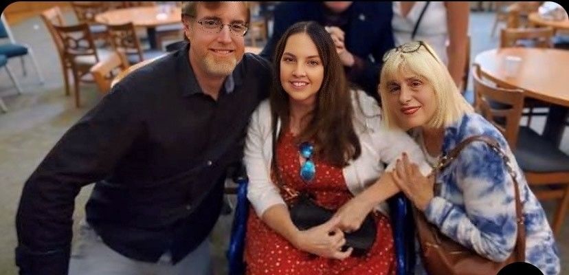 Three people smiling: a man and woman standing next to a woman in a wheelchair, in a restaurant.
