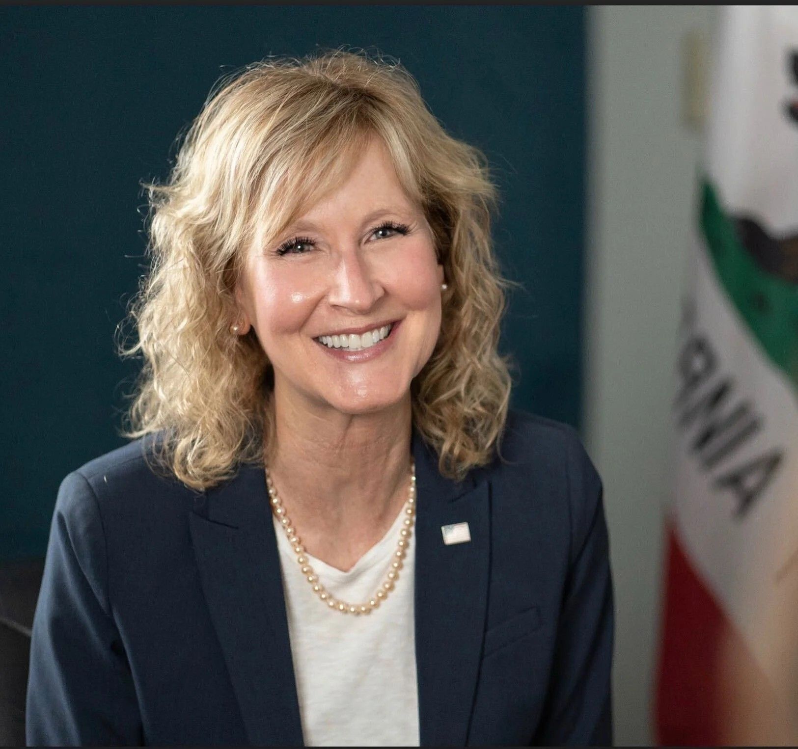 Woman with blonde hair wearing a navy blazer and pearl necklace smiling at the camera, with a California flag in the background.