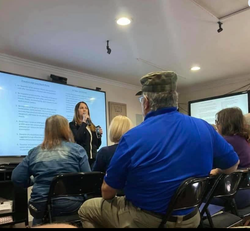 A woman lectures in front of a screen to a seated audience. Several people listen attentively, including a man wearing a camouflage hat.