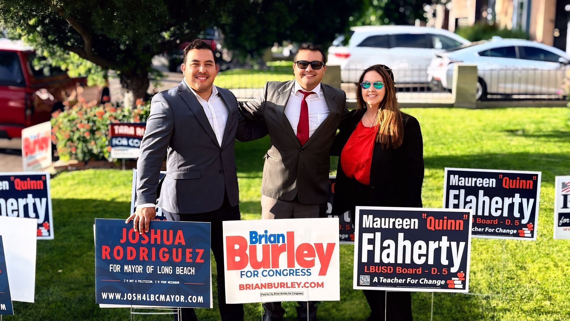 Three people pose on a lawn with campaign signs for Joshua Rodriguez, Burley, and Flaherty.