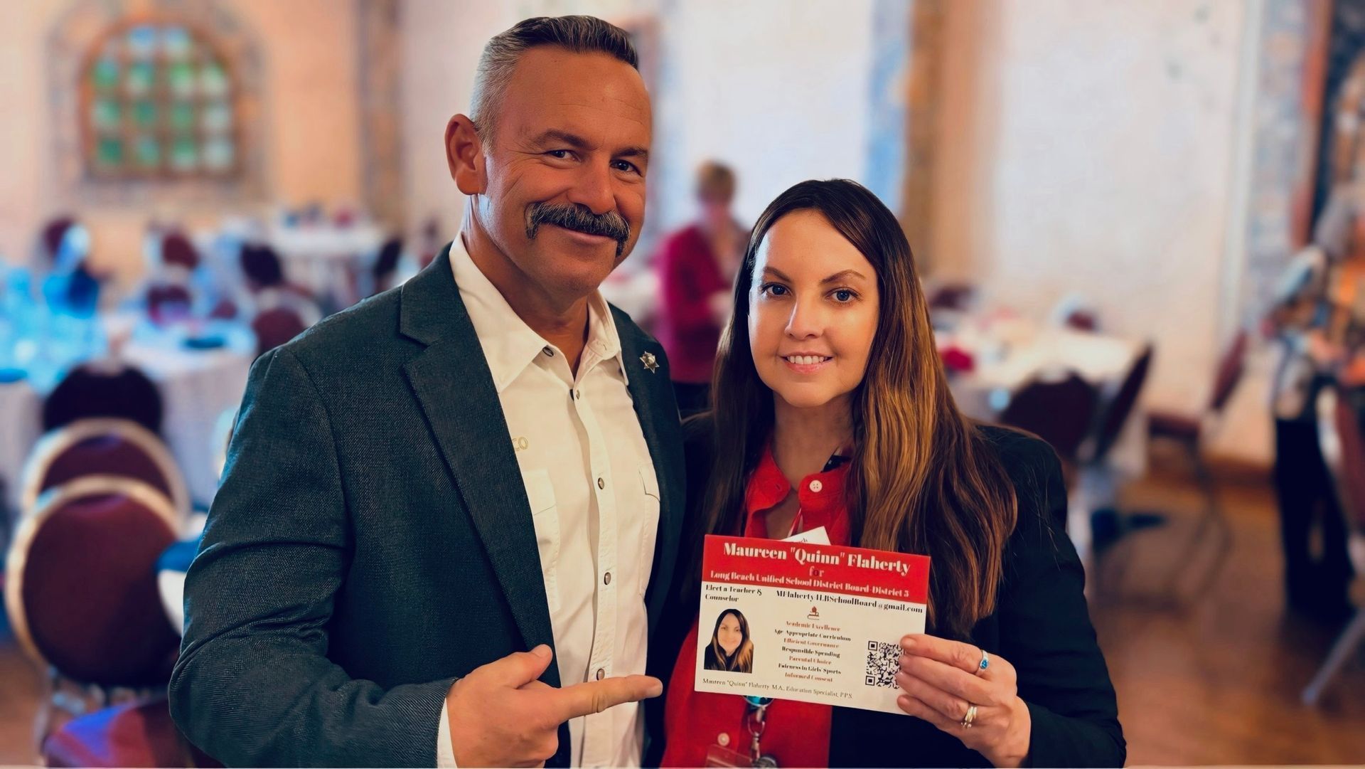 Man in suit points to woman holding ID; both smile in a room with tables and a blurred background.