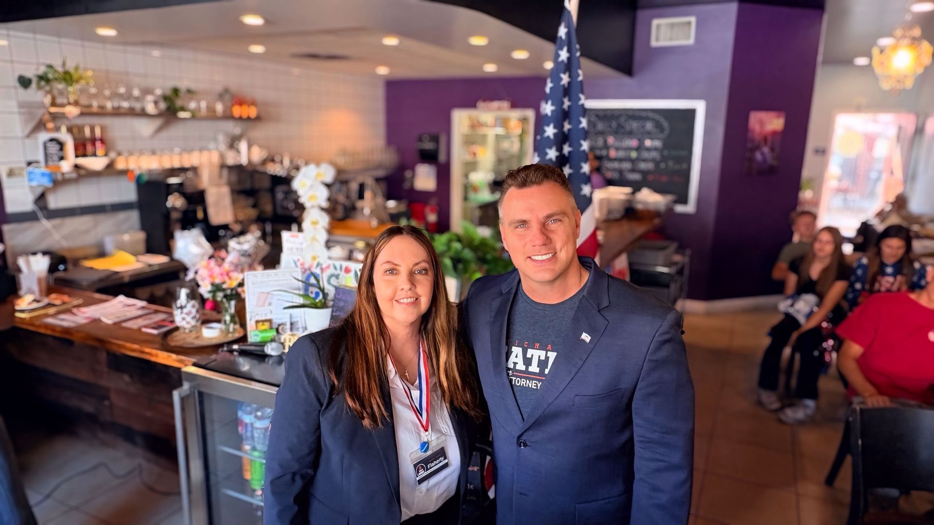 Two people smiling in a café, with a counter, purple walls, and seated patrons in the background.
