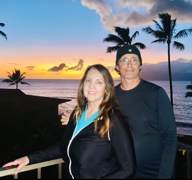 Maureen Flaherty & husband Scott A man and a woman standing on a balcony overlooking the ocean at sunset