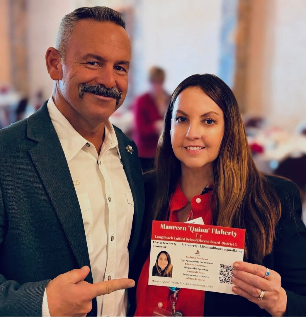 Woman holding microphone and papers, speaking in front of a dark backdrop, wearing a blue shirt and a beige cardigan.