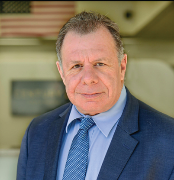 Mike Netter Man in a blue suit and tie, standing outdoors with an American flag in the background. He is looking directly at the camera.