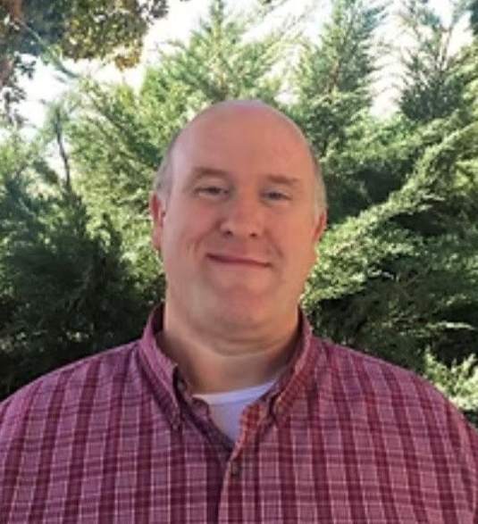 A person in a gray suit and red tie smiling at a desk against a blue background, wearing an American flag lapel pin.