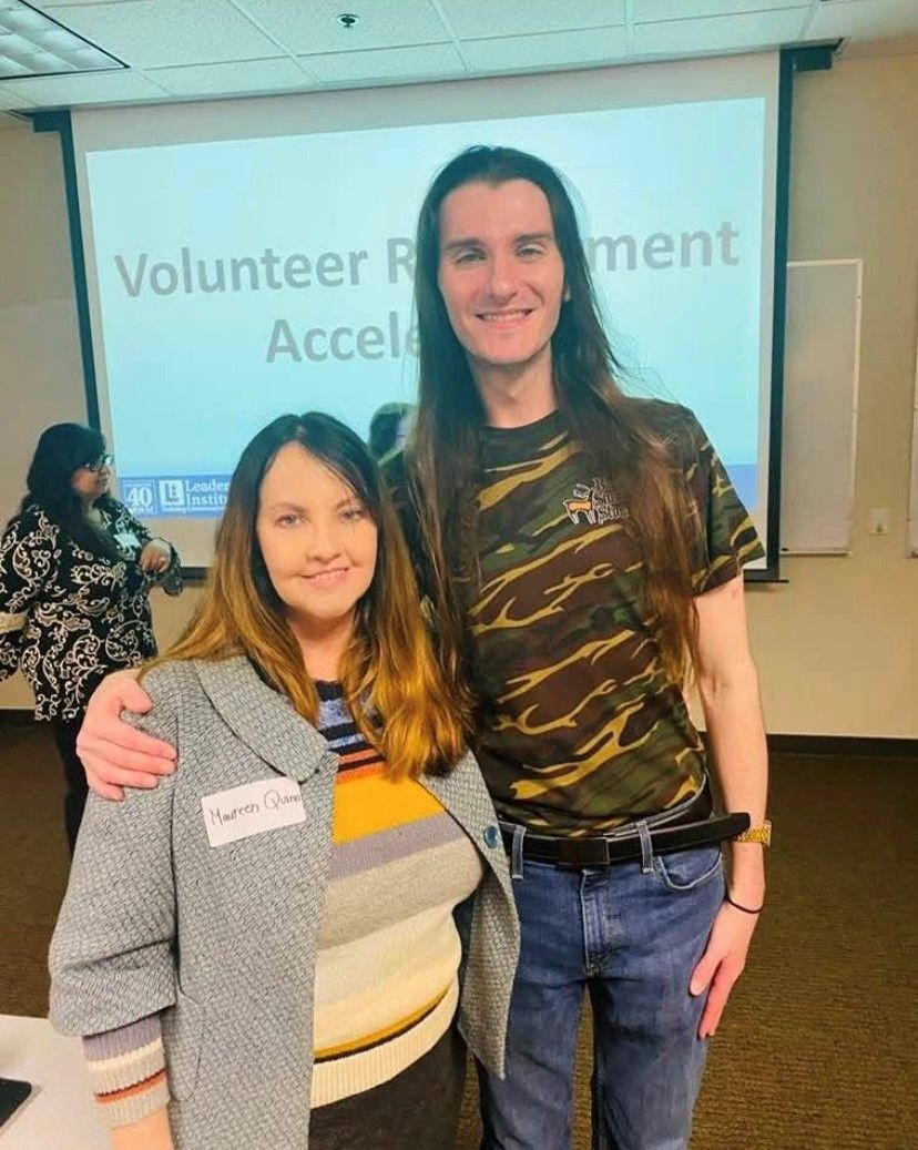 Two people pose for a photo in front of a screen. The woman on the left wears a gray blazer and smiles. The man on the right has long hair, a camouflage shirt, and his arm around her.