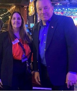 Woman with red top and man in blazer pose for a photo. Indoor setting, party lights in background.