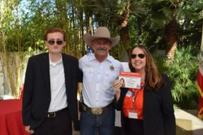 A man in a cowboy hat and sheriff's uniform poses with a young man and woman holding a card outdoors.