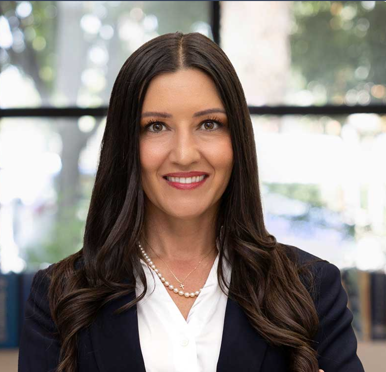 Leslie Manookian Woman with long brown hair, wearing a navy suit and pearl necklace, smiling, in an office setting.