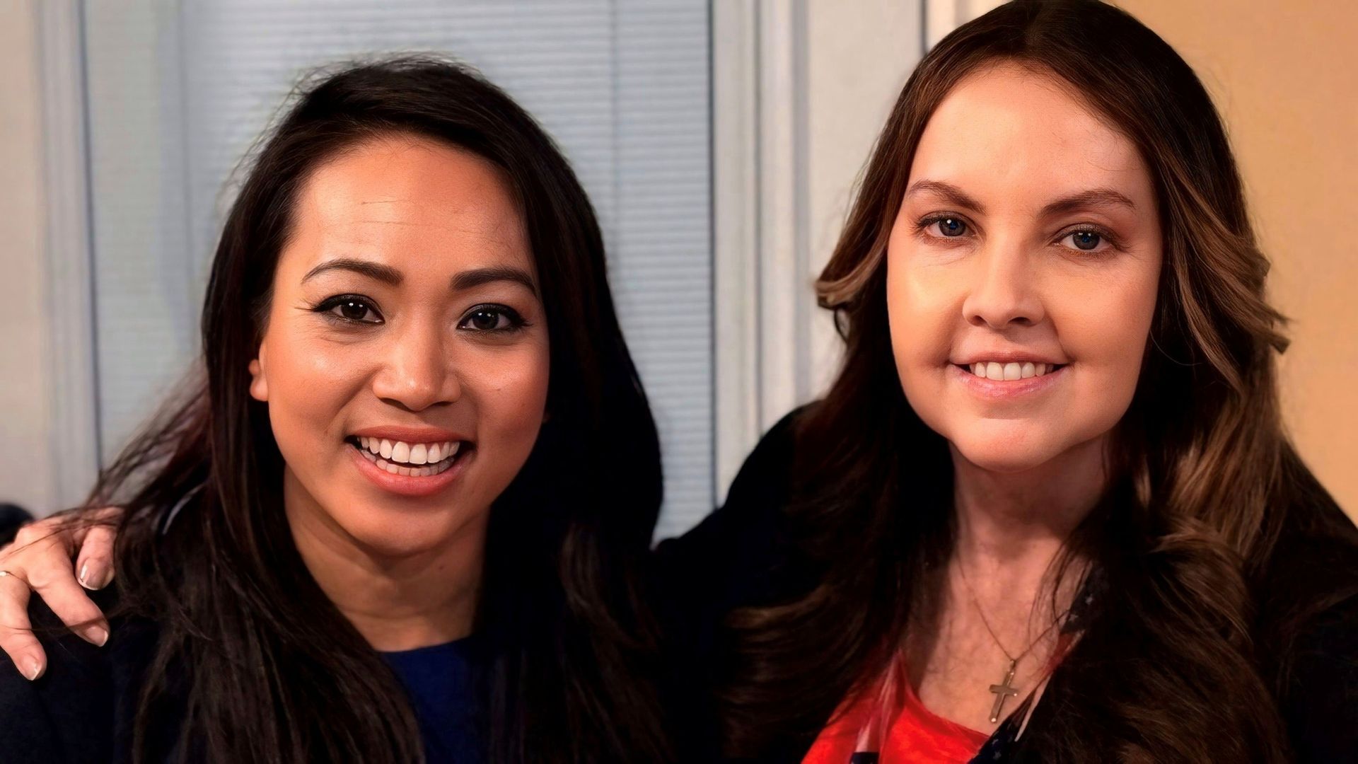 Two women smiling, one has arm around the other. Interior setting, one in blue, one in red.