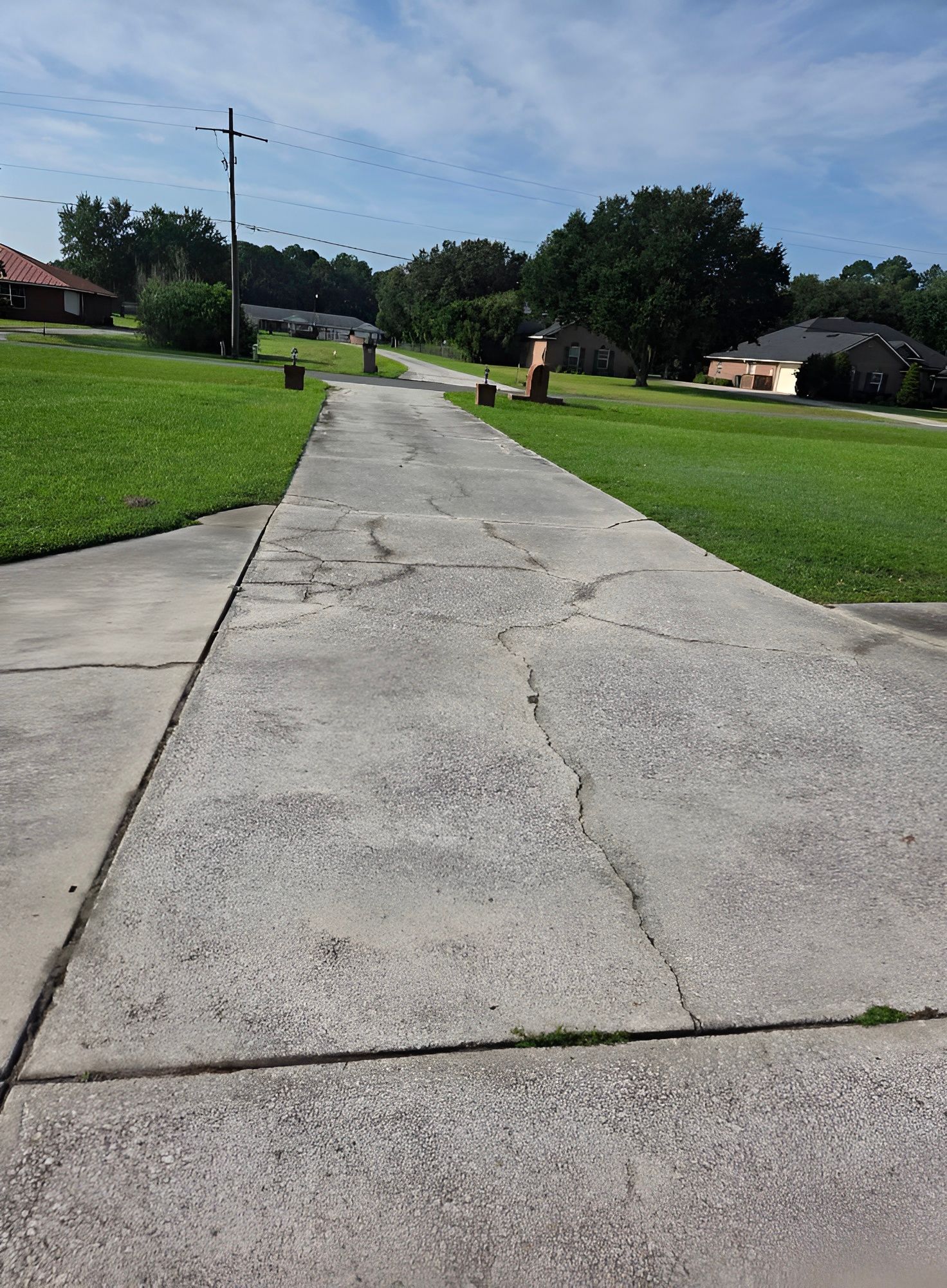 Cracked concrete driveway leading toward houses on a sunny day. Green grass on either side.