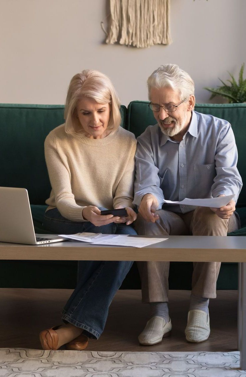 An elderly couple is sitting on a couch looking at a laptop and a cell phone.