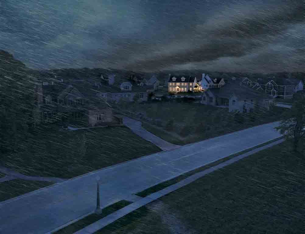 Une rue de quartier résidentiel au crépuscule, avec une maison illuminée parmi plusieurs autres sous un ciel sombre et nuageux.