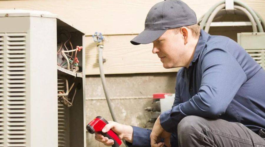 A man is kneeling down next to an air conditioner while holding a flashlight.