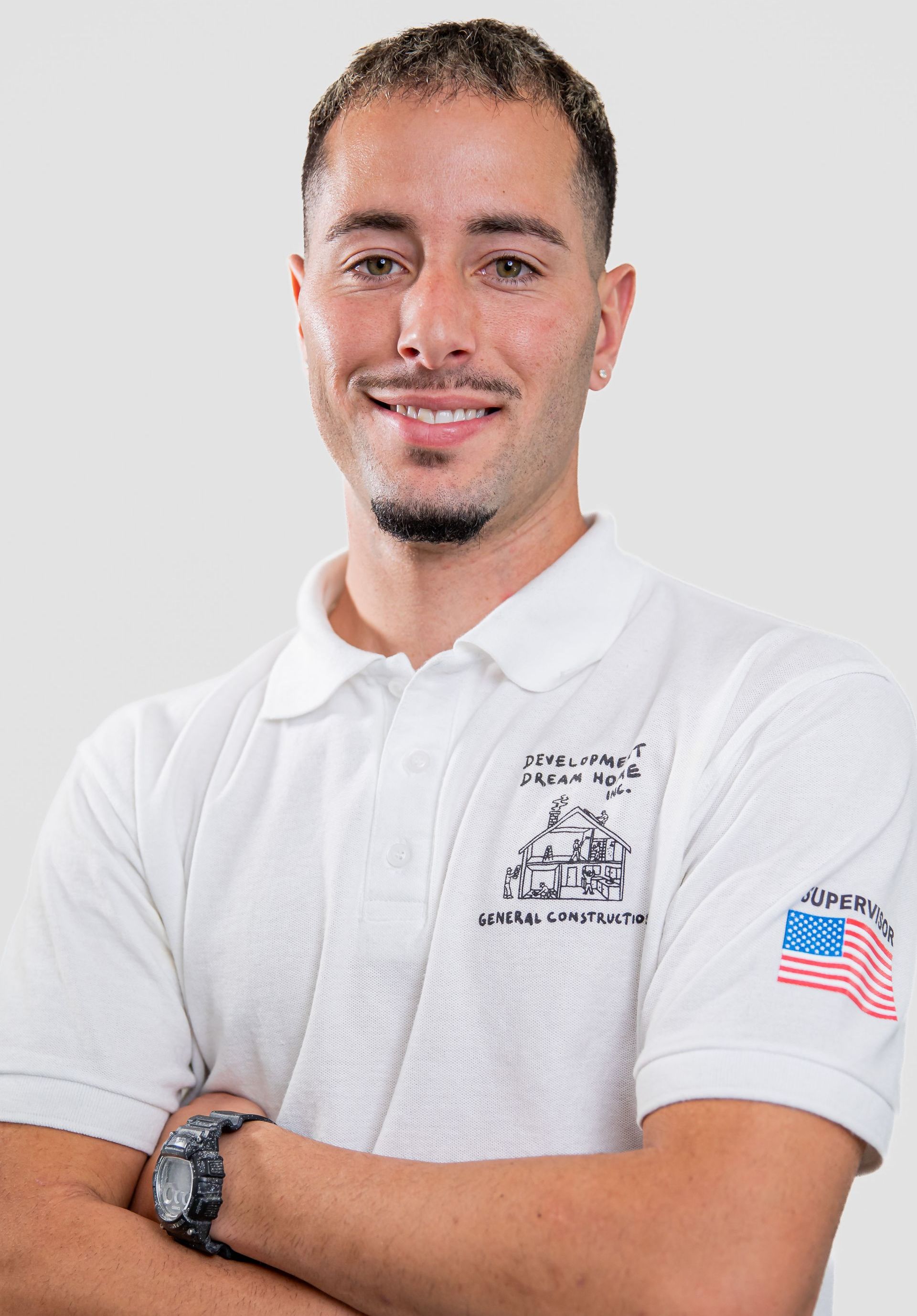 A man wearing a white polo shirt and a watch is smiling with his arms crossed.