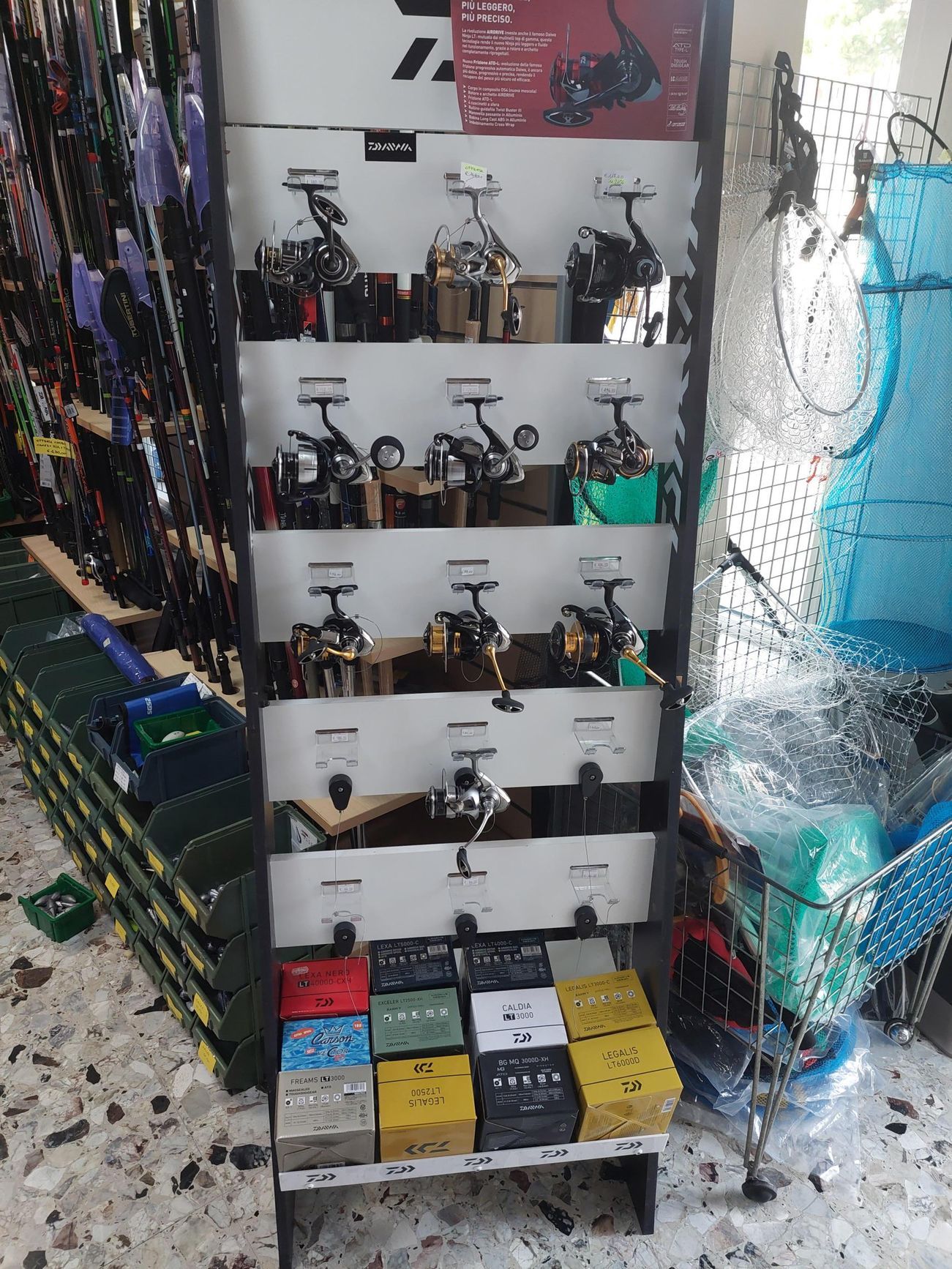 Mulinelli da pesca esposti in un negozio. Scaffali con mulinelli, scatole sottostanti. Retini e canne da pesca sullo sfondo.