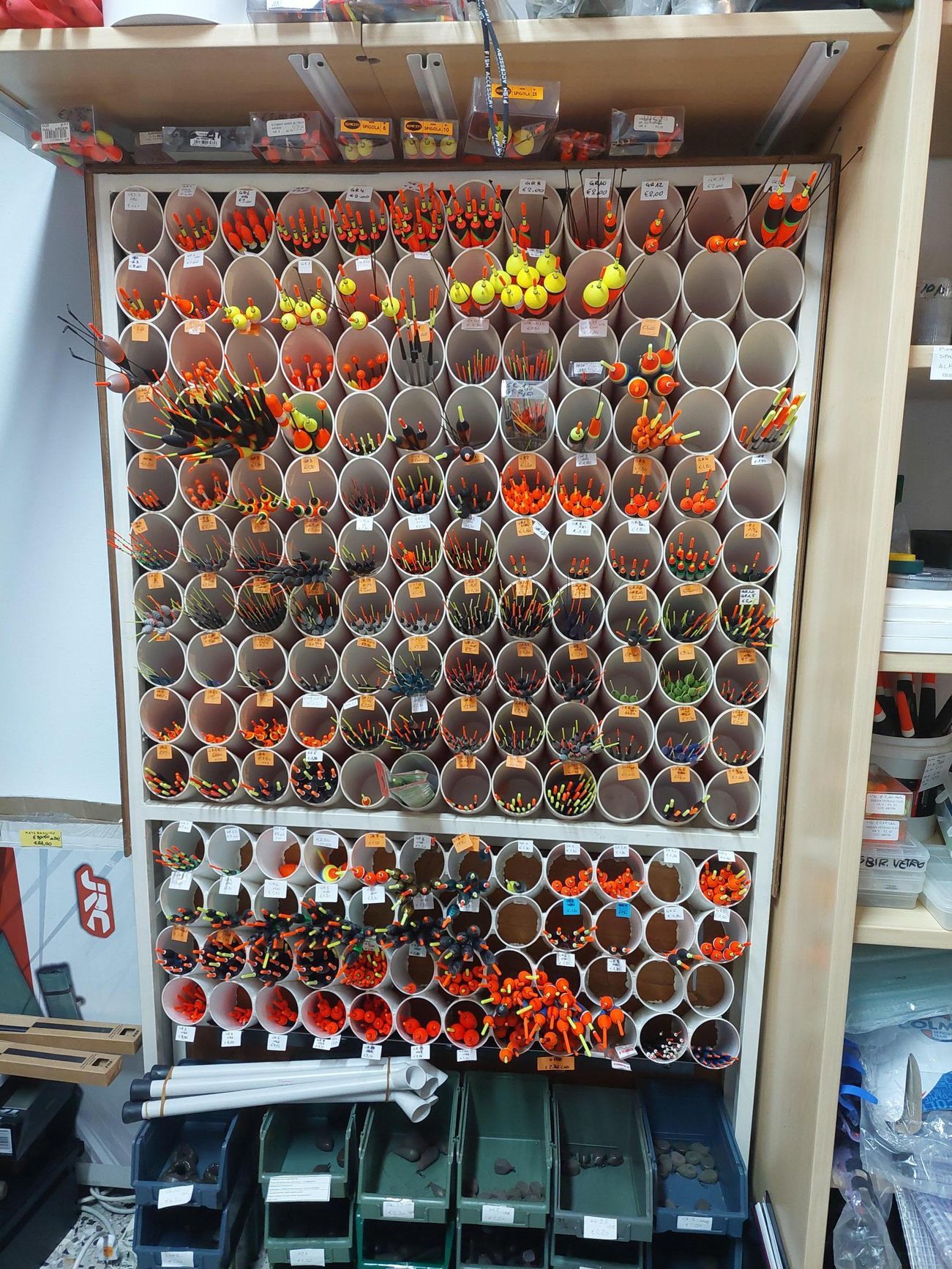 Una parete espositiva di galleggianti da pesca disposti in tubi bianchi. Sono visibili vari colori.