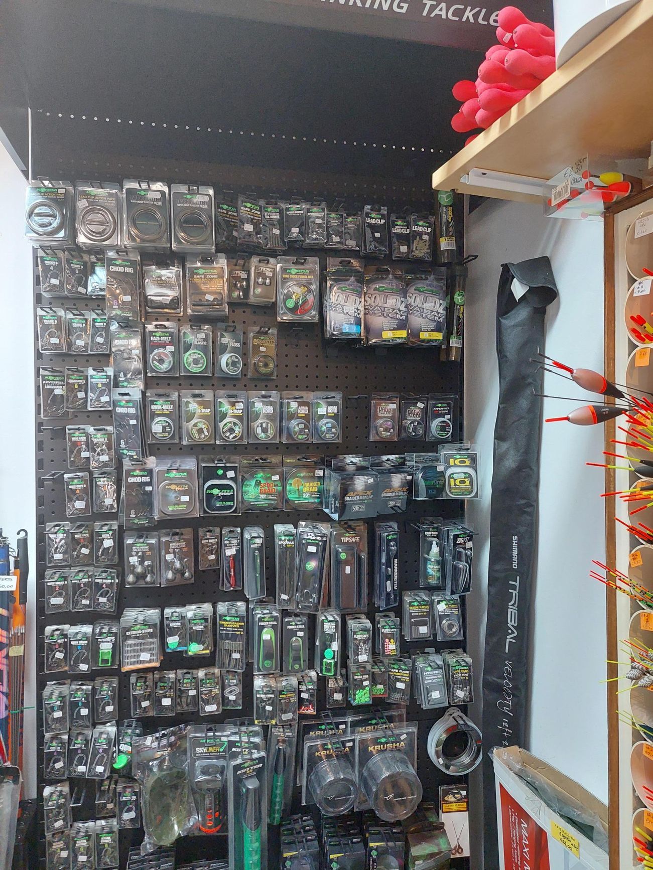 Espositore di attrezzatura da pesca in un negozio, organizzato su un pannello forato con vari articoli confezionati. Appesa una custodia per canna da pesca.