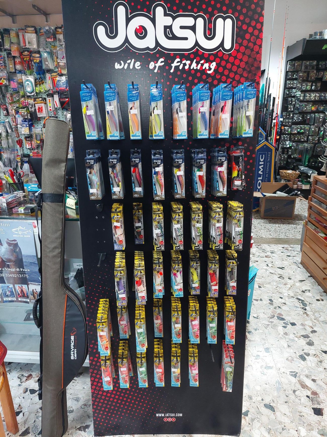 Esposizione di esche da pesca in un negozio. Insegna del marchio Jatsui. Numerose confezioni di esche su pannello nero.