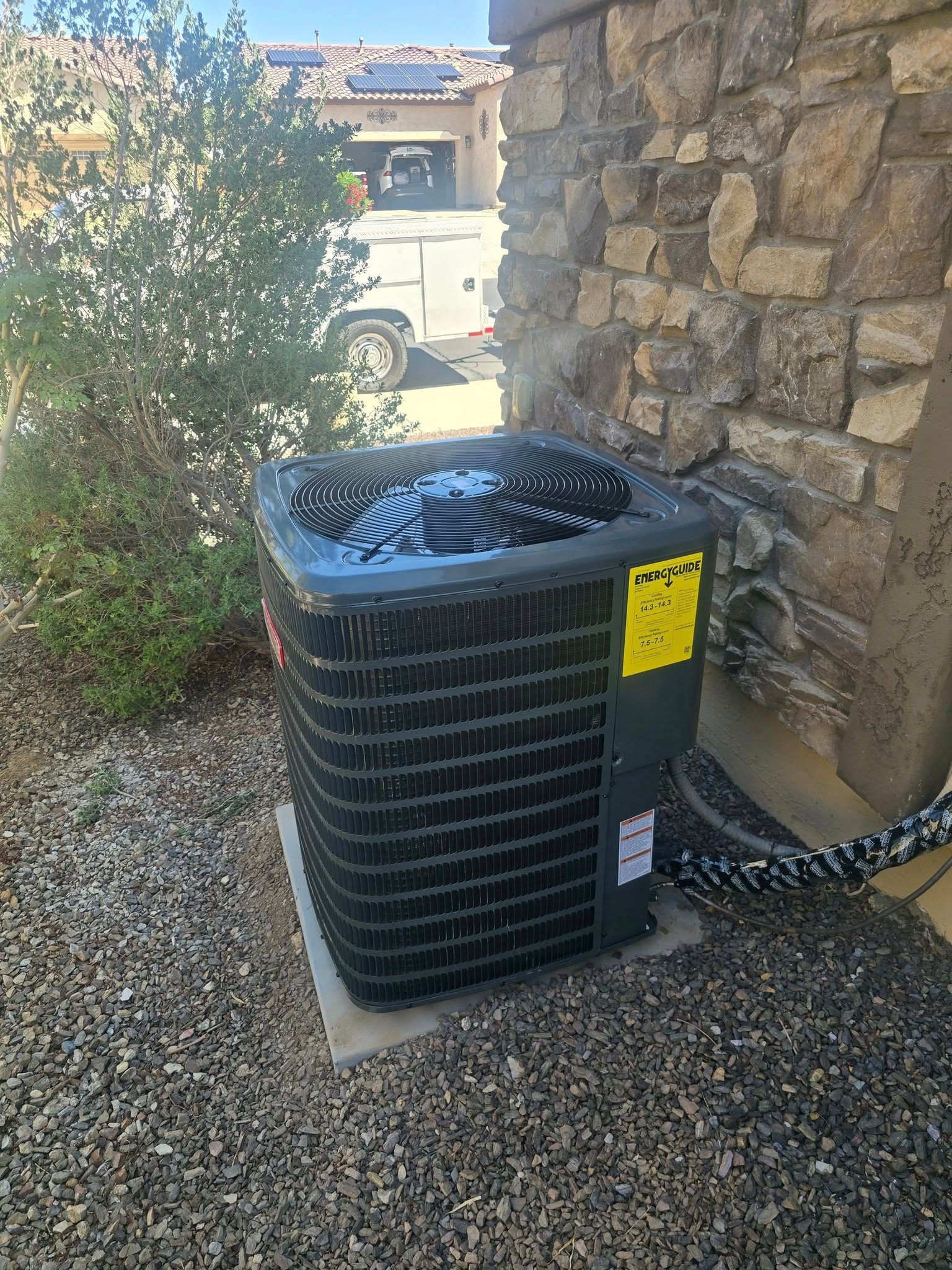 AC unit next to a stone wall on a gravel surface, with a bush and part of a vehicle in the background.