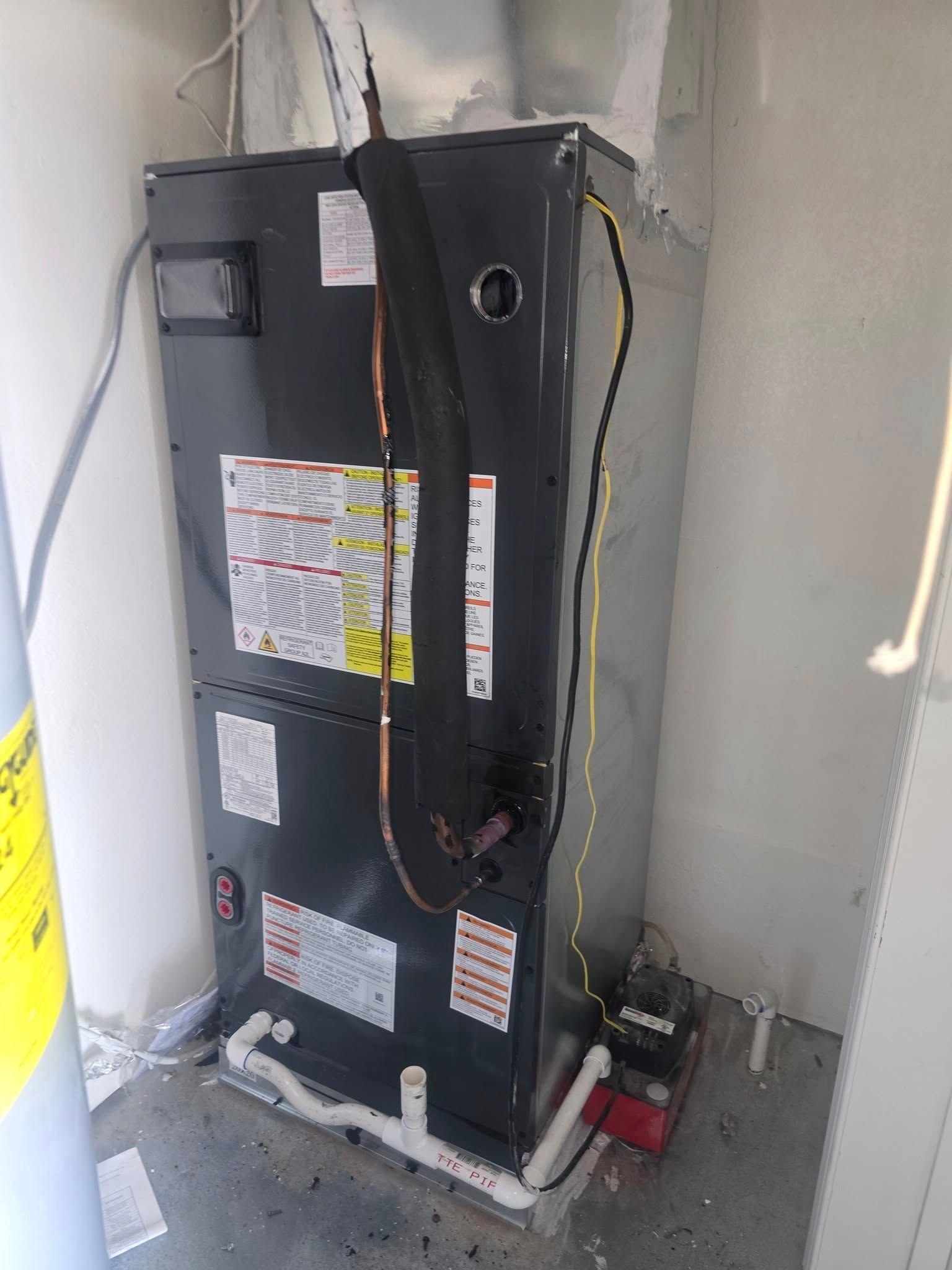 An air handler unit in a utility closet, against a white wall, with pipes and wires connected to it.