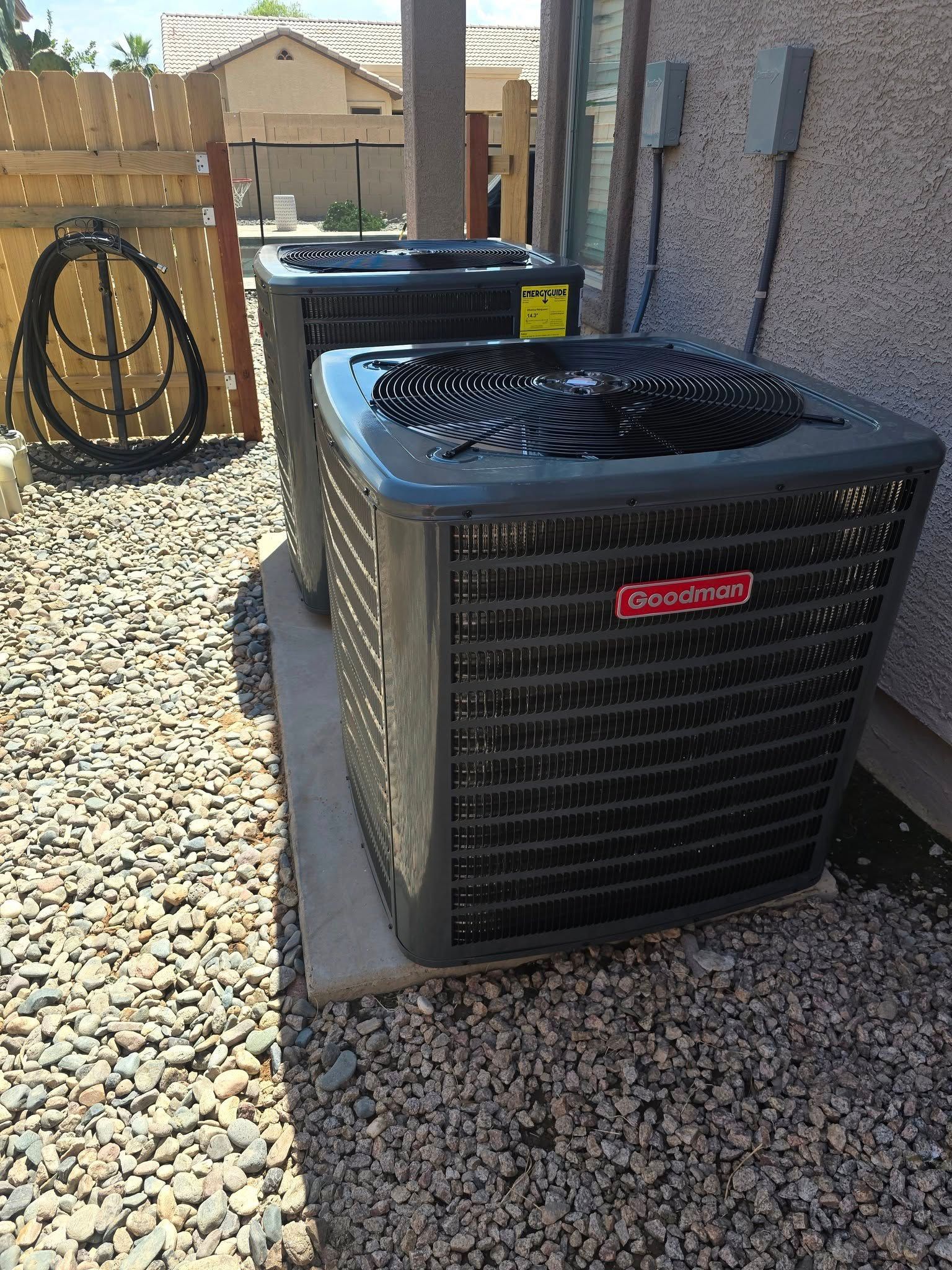 Two Goodman AC units on a concrete pad, in a gravel area next to a building.