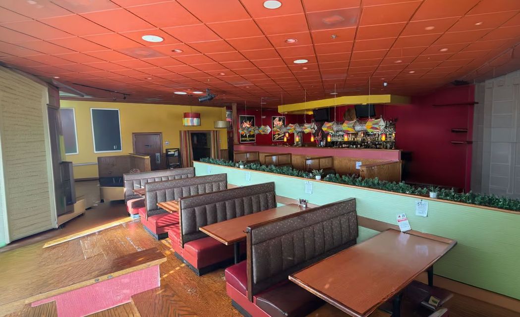 Empty restaurant dining area with booths and tables, orange ceiling, and a bar stocked with bottles.