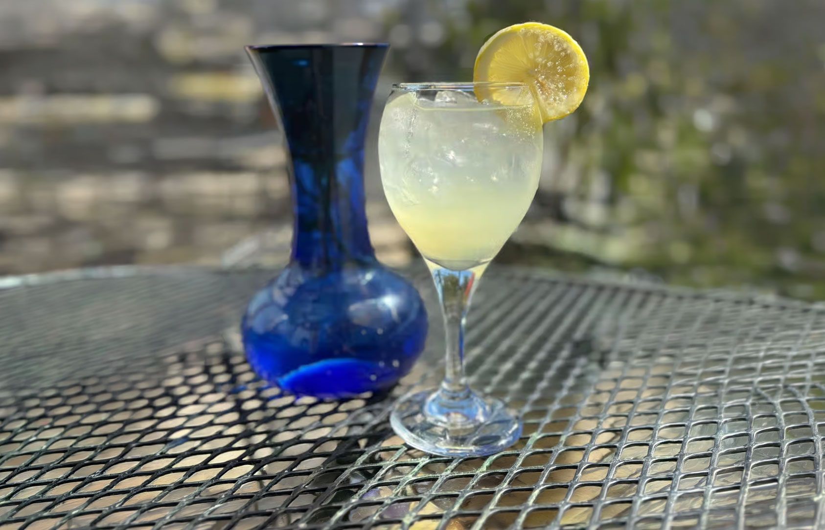 Blue vase beside a pale yellow drink with lemon slice on a metal patio table