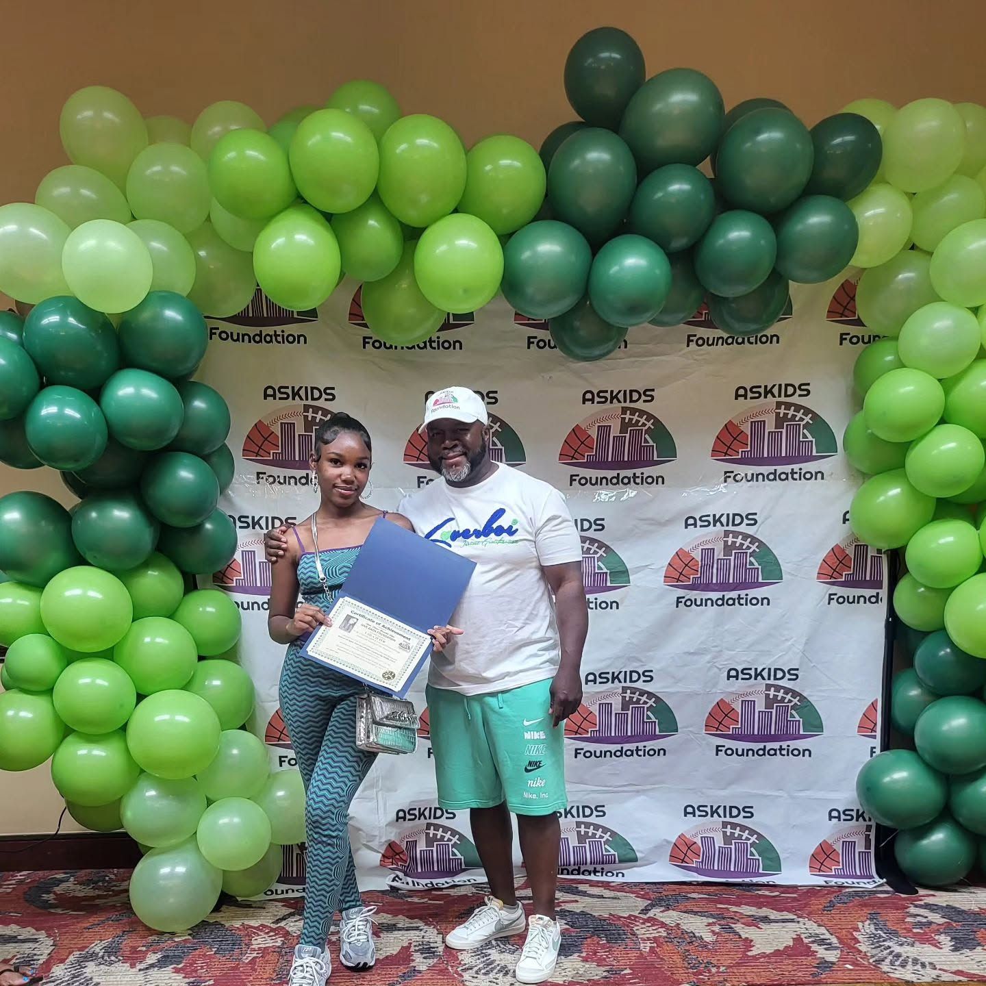 Two people posing in front of a green balloon photo backdrop at an event