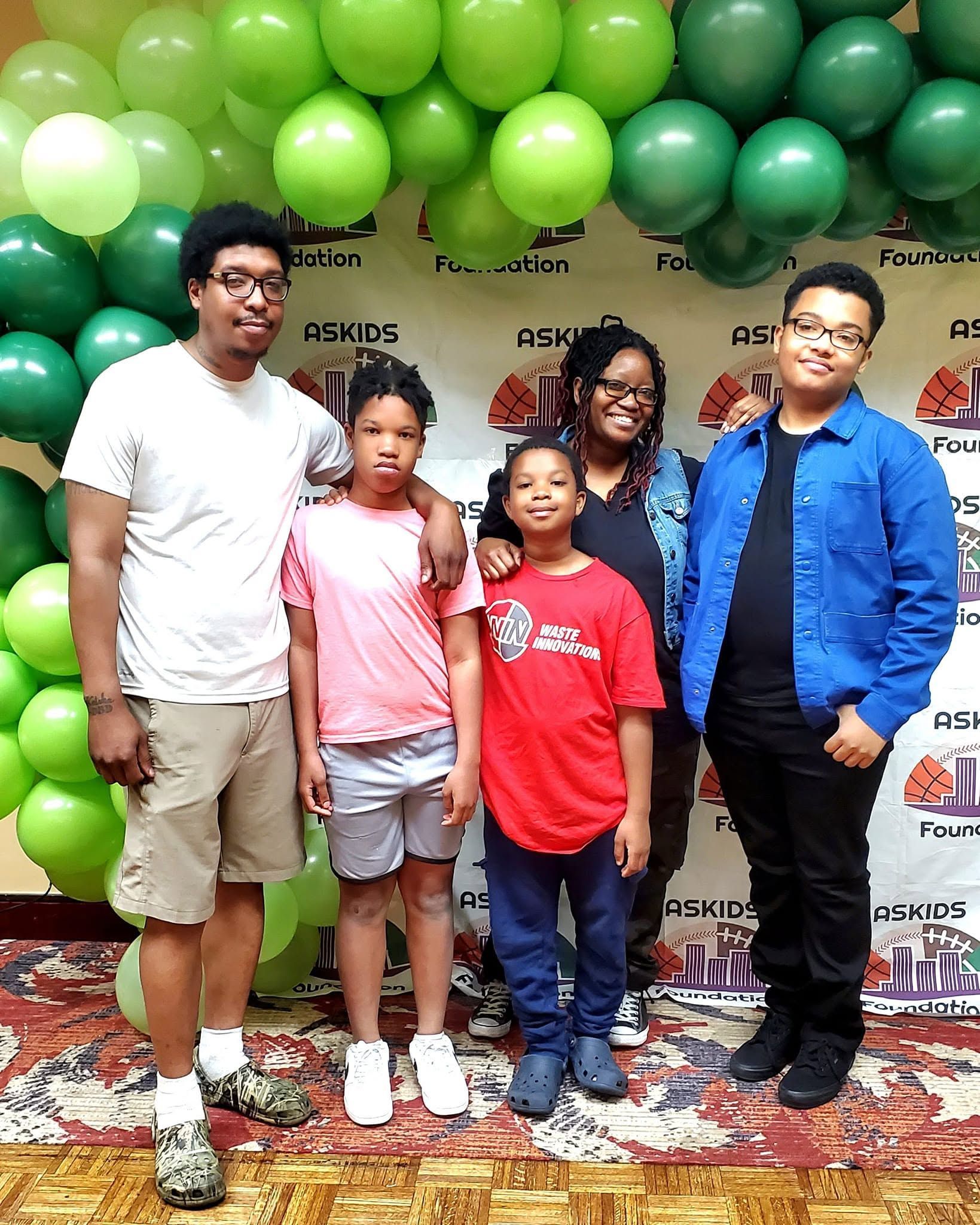 Four people posing under green balloons at a party backdrop, wearing casual colorful outfits.