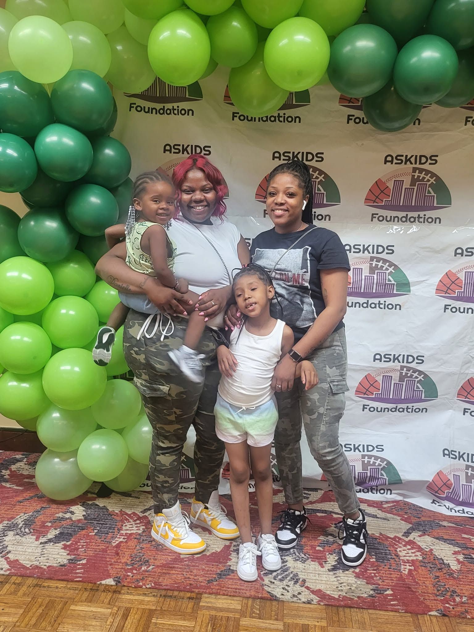 Three people posing in front of a green balloon backdrop at an event, two adults holding a child.