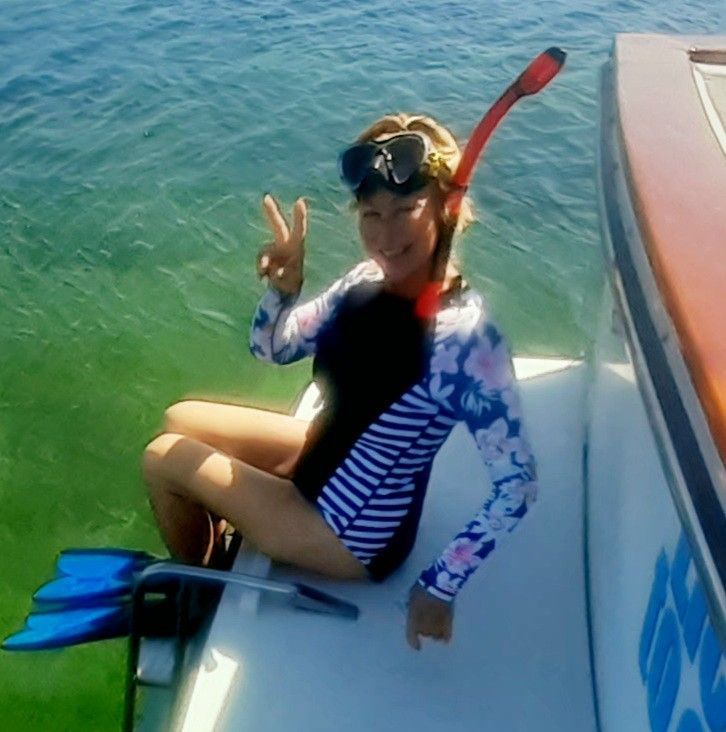 Woman in snorkeling gear, seated on a boat, gives a peace sign, water and blue fins visible.