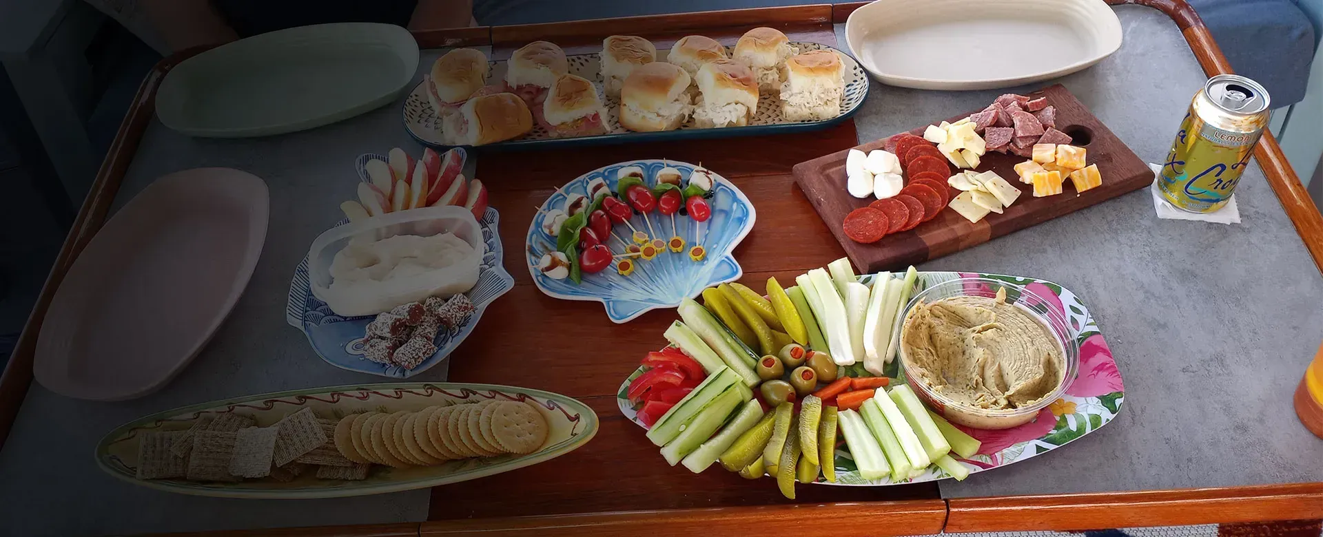A wooden table with various snack platters: crackers, dips, cheese, cured meats, veggies, and baked rolls.
