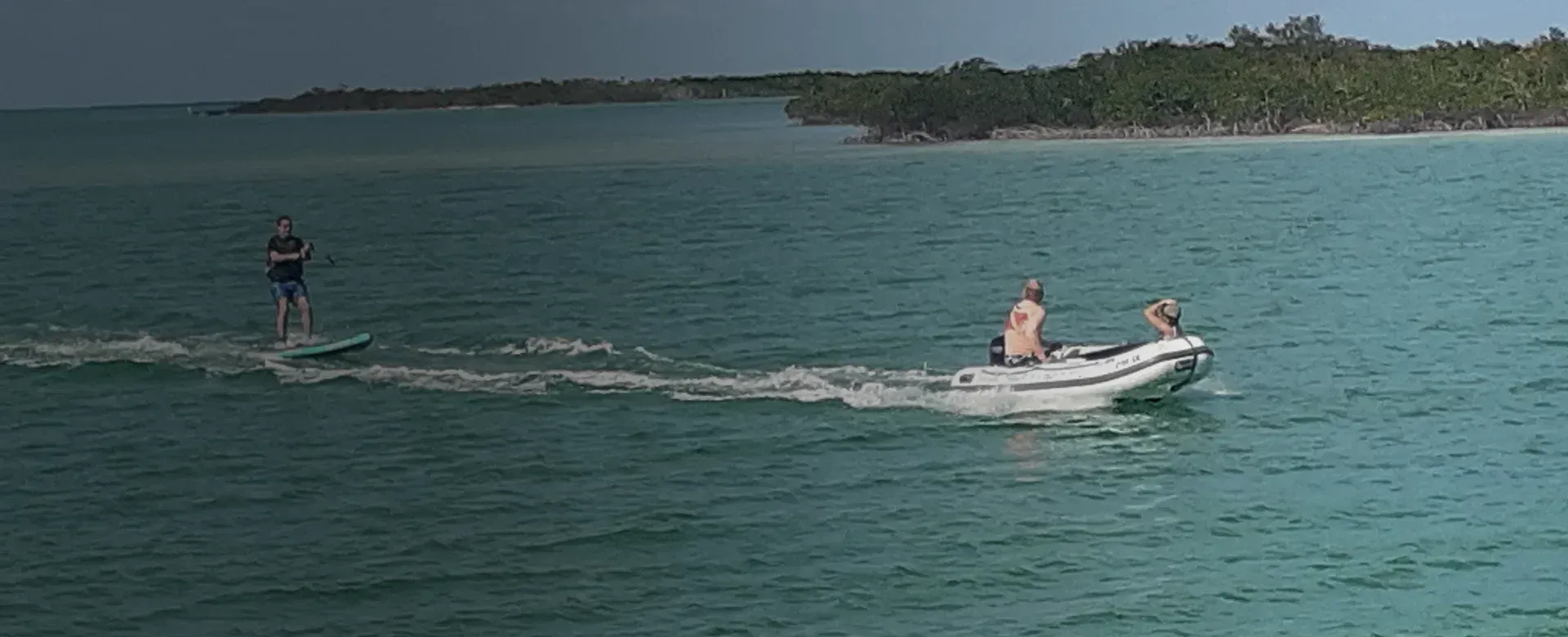 A person on a hydrofoil surfboard is being towed by a motorboat in turquoise water near a shoreline.