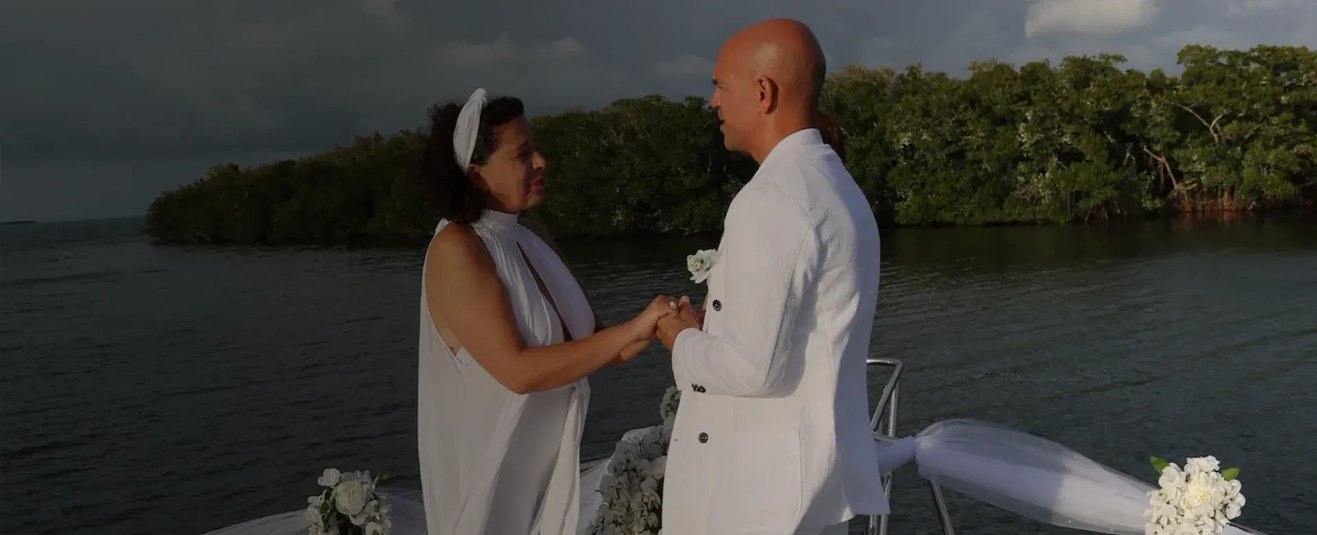 Couple holding hands on a dock for a waterside wedding. White decor and a wooded backdrop.