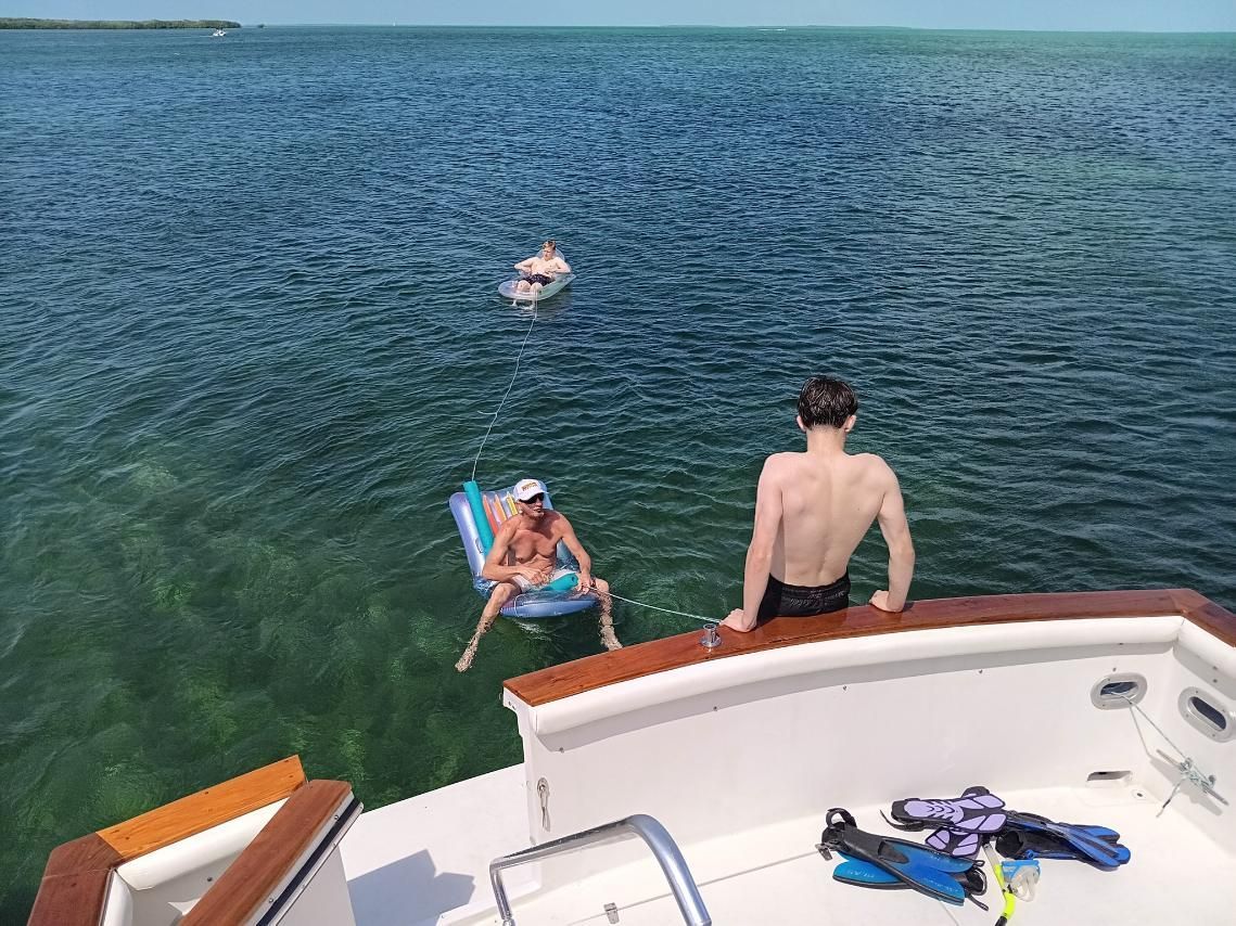 People enjoying water near a boat; one floats on an inflatable, another sits on the boat edge, and one swims.