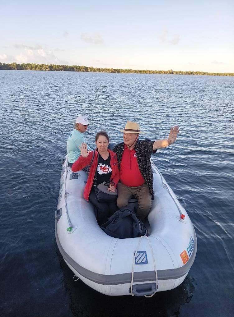 Three people wave from a white inflatable boat on a calm body of water.