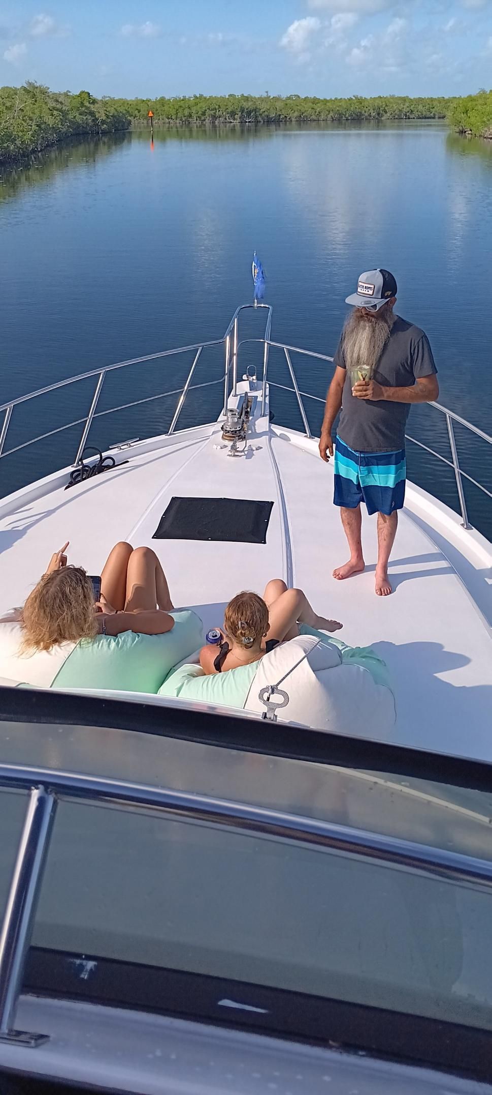 People on a boat sailing on a river. One man standing, two people sunbathing. Lush greenery in the background.