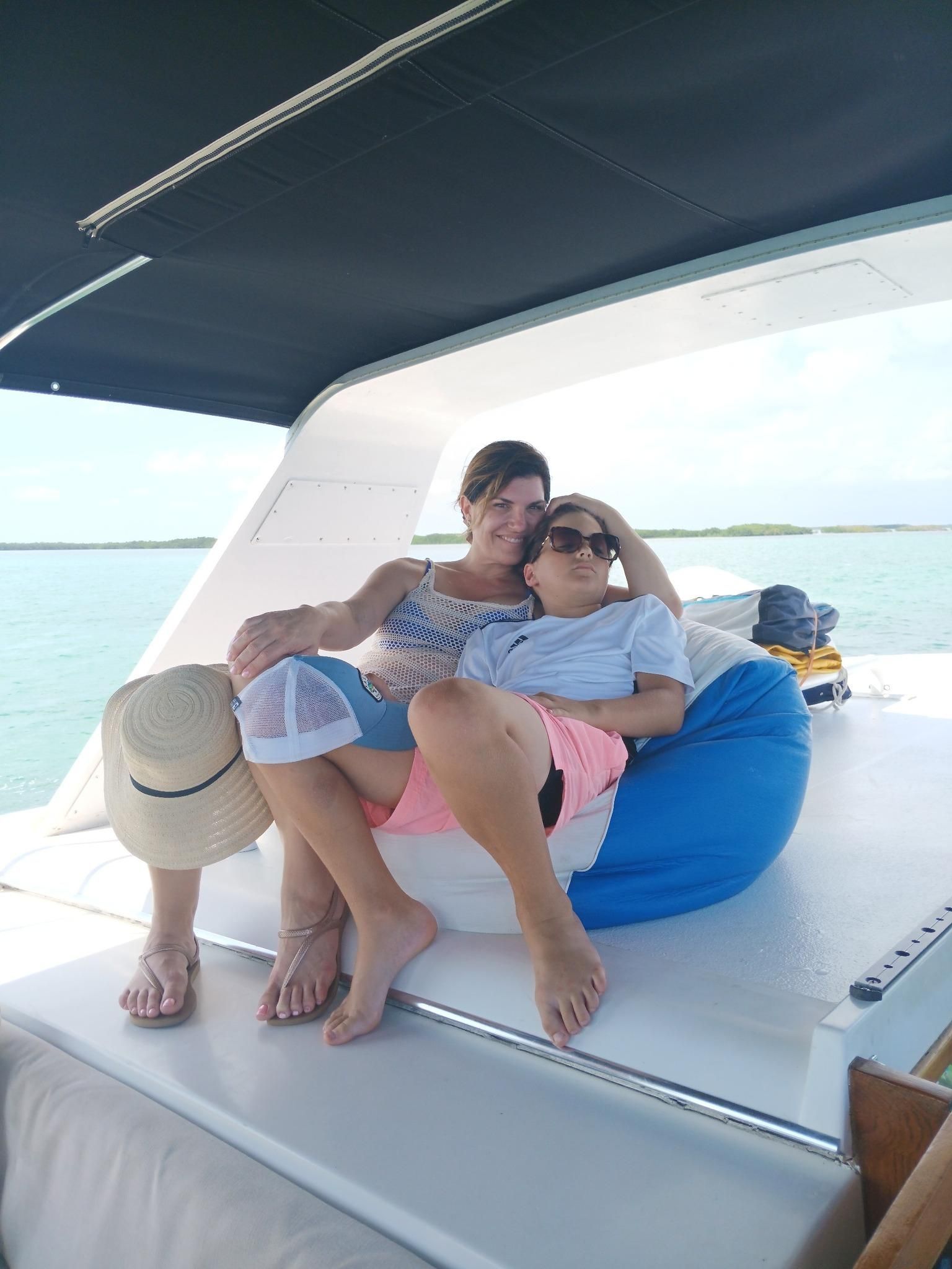 Two women relax on a boat, one with her arm around the other. They are sitting on a beanbag and smiling, with blue water in the background.