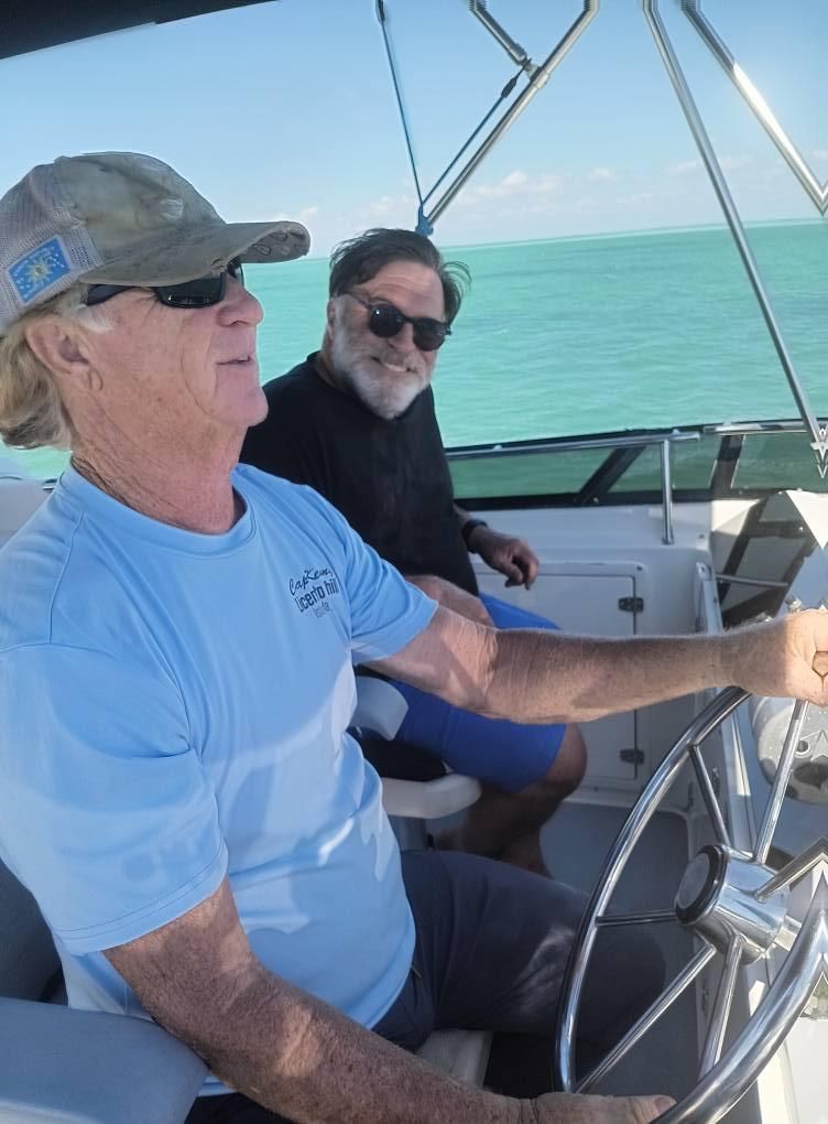 Two men on a boat; one steers, wearing a cap and sunglasses. Turquoise water in the background.