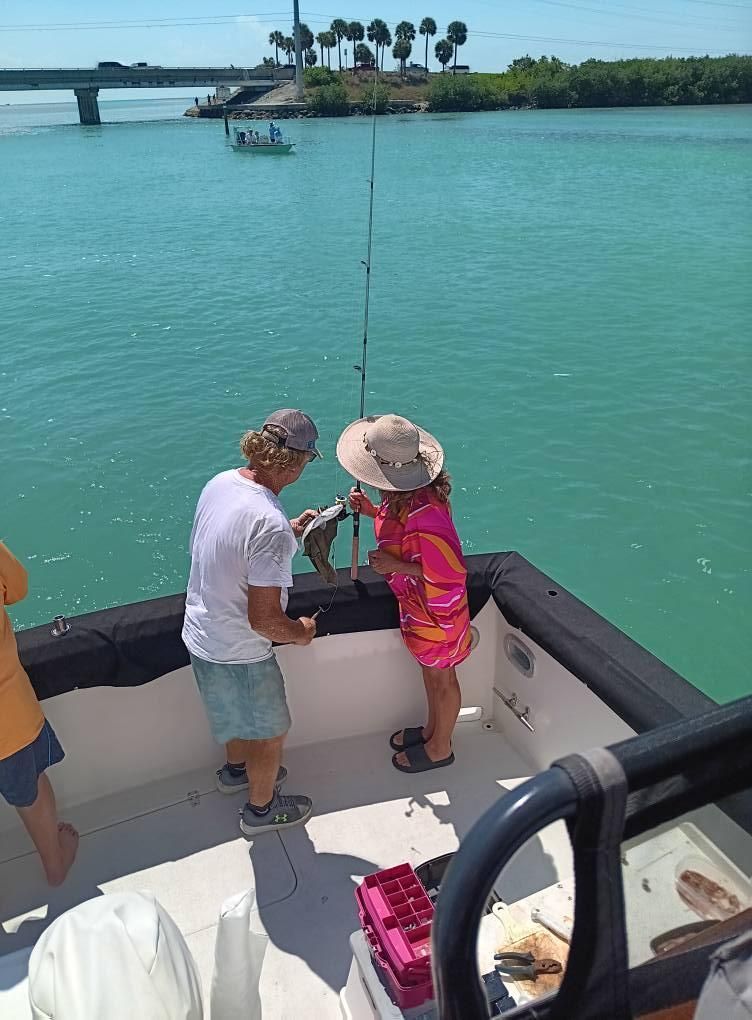 Two people on a boat, one holding a fish near water. Bridge and other boat in background.