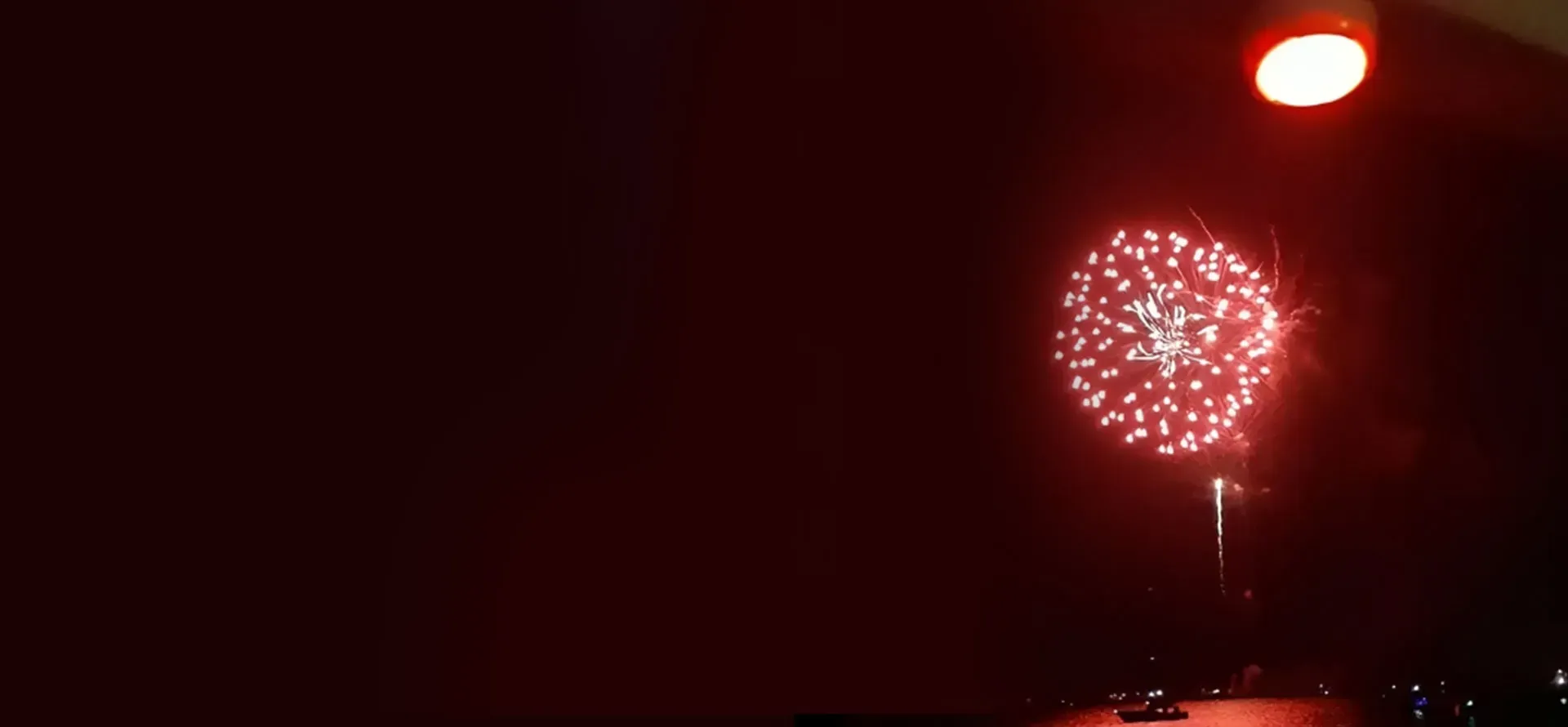 Fireworks burst in a red sky, with a bright light in the corner.
