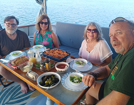 Four people eating a meal at a table on a boat, with the ocean visible in the background.