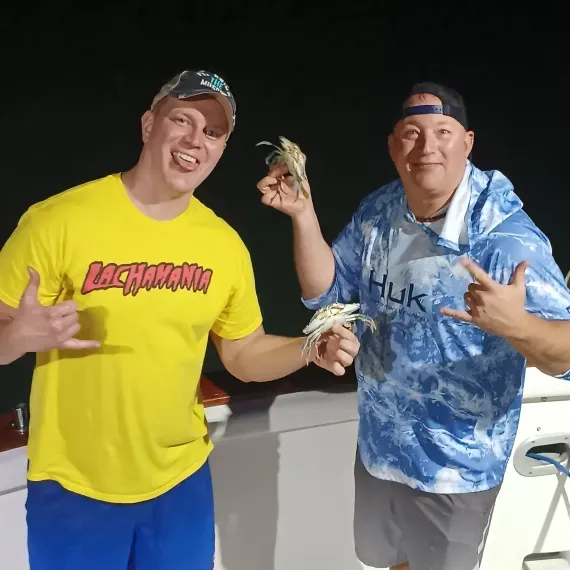 Two men on a boat at night holding up shrimp, making hand gestures. One wears yellow, the other blue.
