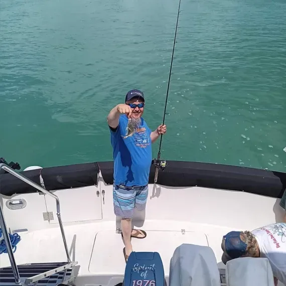 Man on boat, holding up a small fish. Blue water, sunny day.