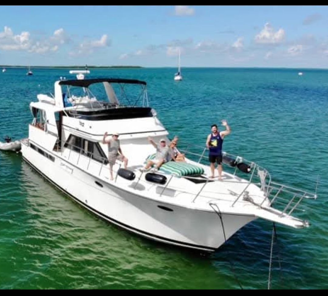 People waving on a white motor yacht in turquoise water. Clear sky and small boats visible.