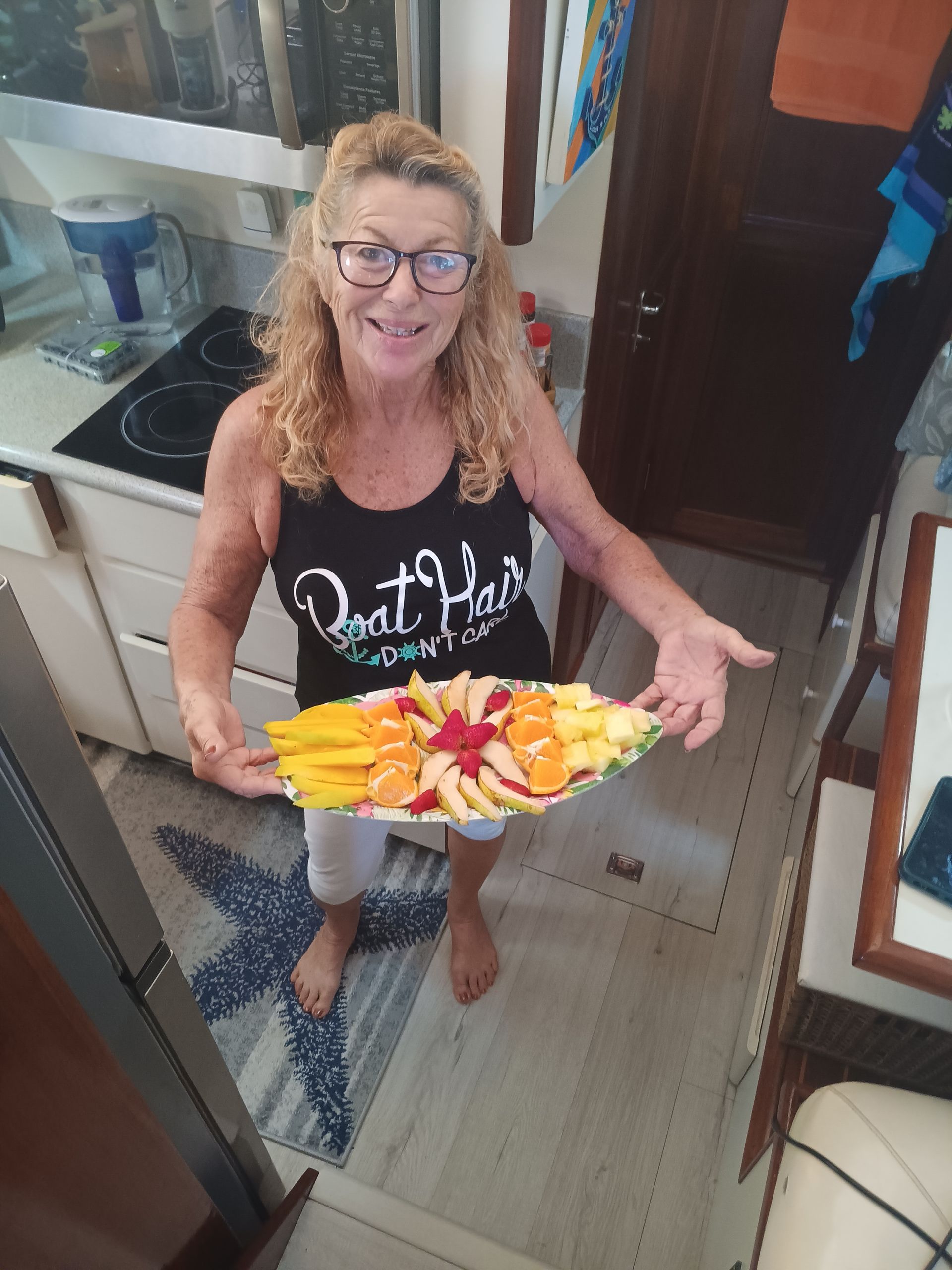 Woman holding a colorful fruit platter in a boat's galley; smiling, wearing glasses and a tank top.